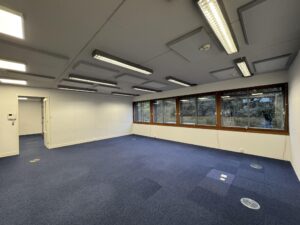 Empty office space with blue carpet flooring, fluorescent ceiling lights, and large windows at De Ruijterkade.